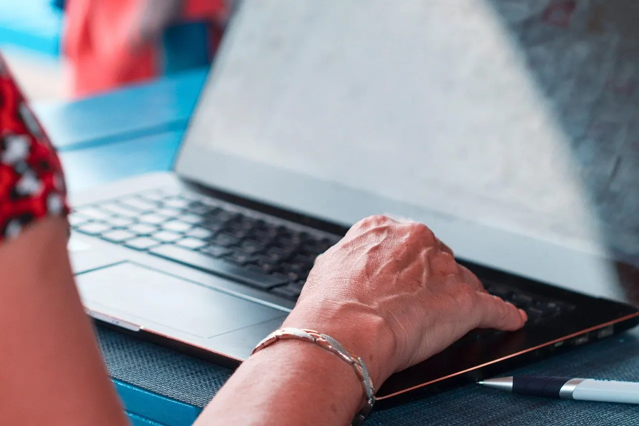 Hand mit Armband tippt auf einer Laptop-Tastatur, daneben ein Stift auf dunklem Untergrund.