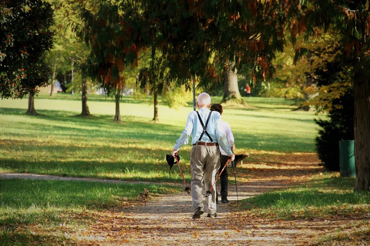 Älterer Mann mit grauem Haar und Hosenträgern geht mit einer Frau und Stöcken auf einem Parkweg zwischen Bäumen.