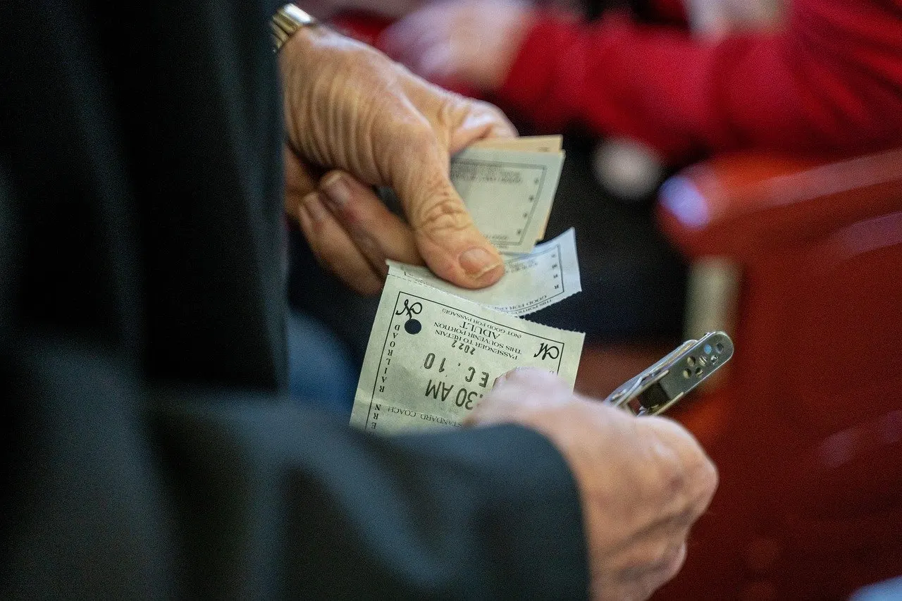 Ältere Hände halten Tickets und Geld, im Hintergrund unscharfe rote Sitzreihen.