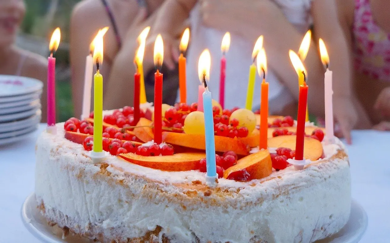 Torte mit Sahne, Früchten und zahlreichen brennenden bunten Kerzen auf einem Tisch im Freien.