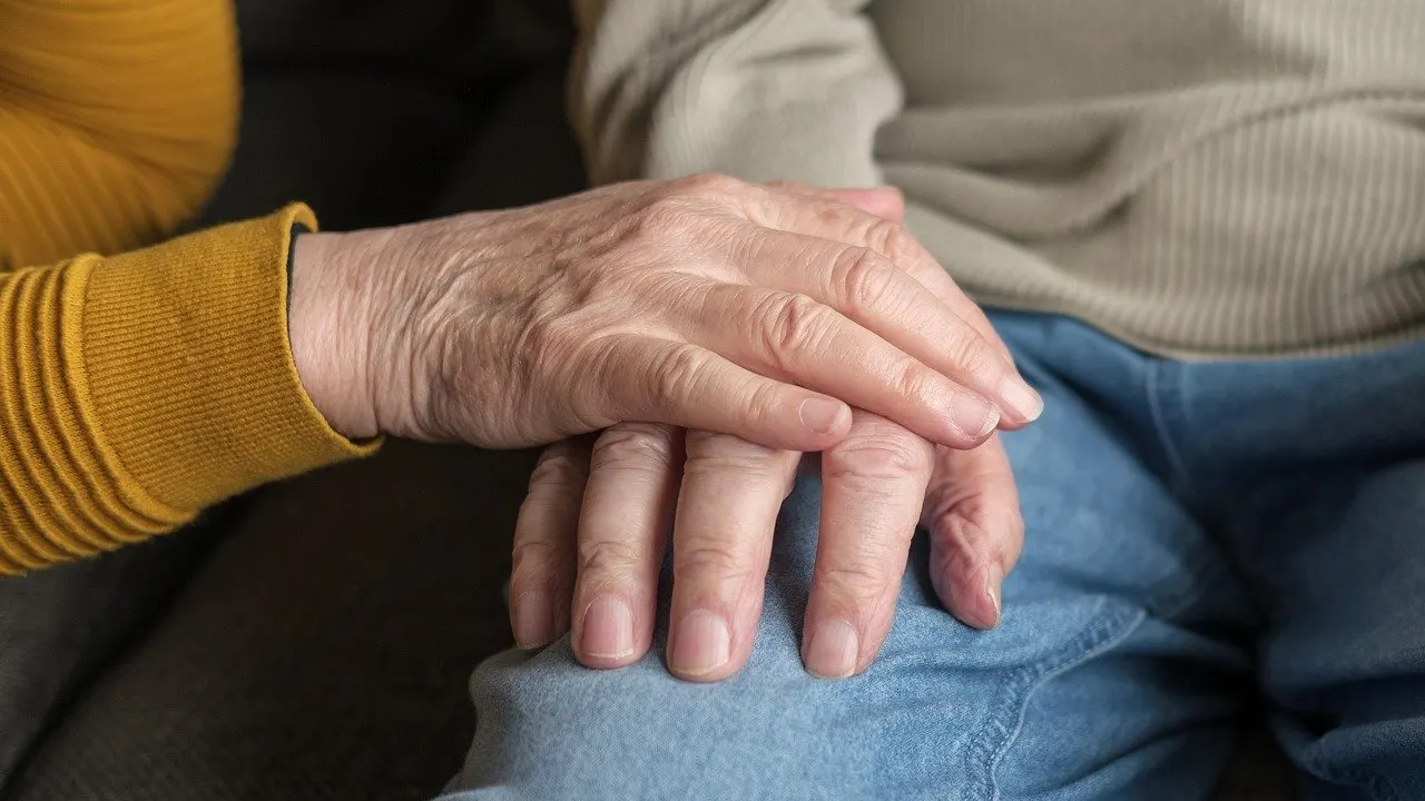 Zwei Hände liegen übereinander auf einem blauen Stoff; die untere Hand ist faltig, die obere gehört zu einem Arm in gelbem Pullover.