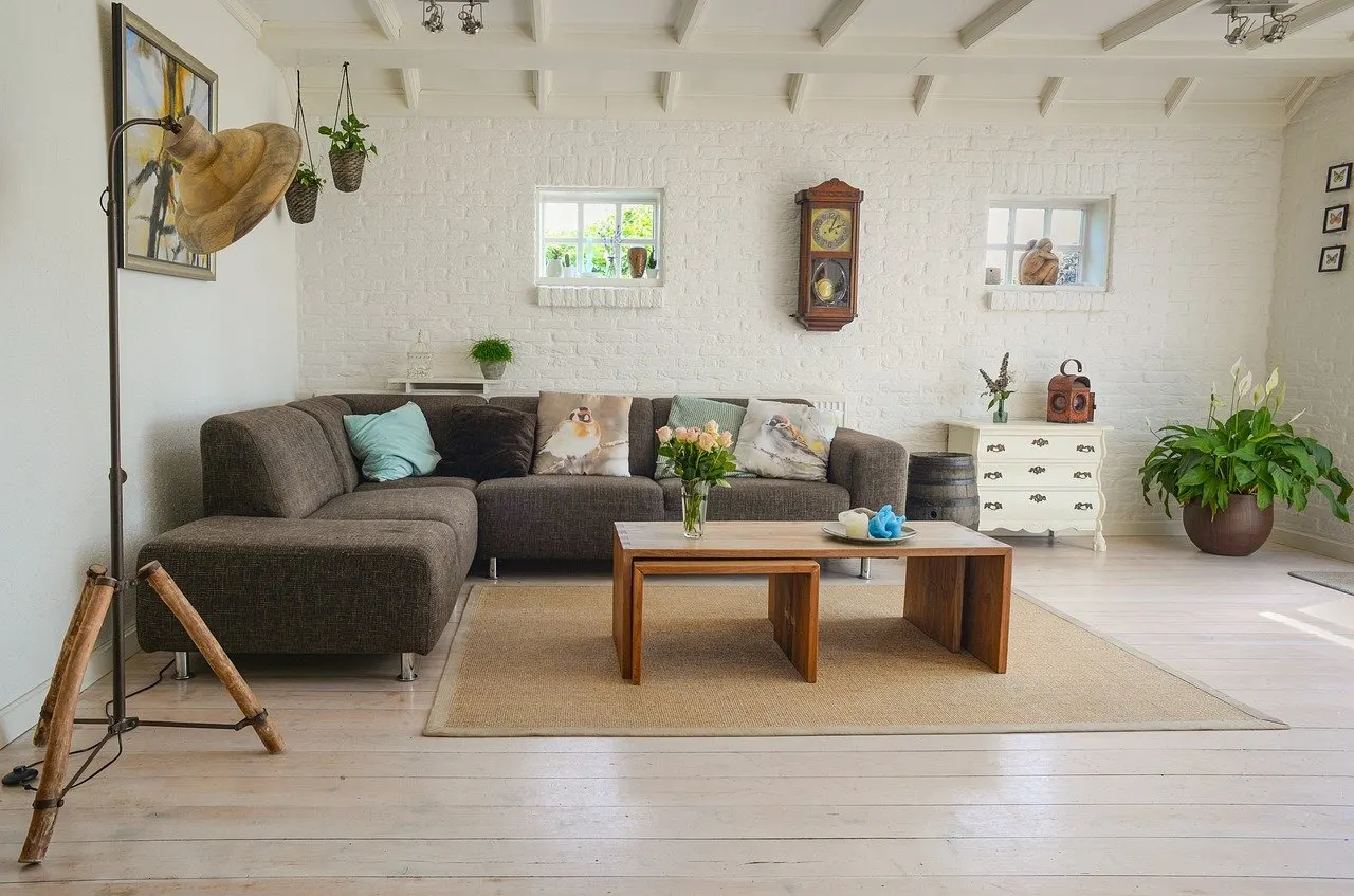 Helles Wohnzimmer mit grauem Ecksofa, Holztisch, Kissen, Wanduhr und Pflanzen auf hellem Holzboden.